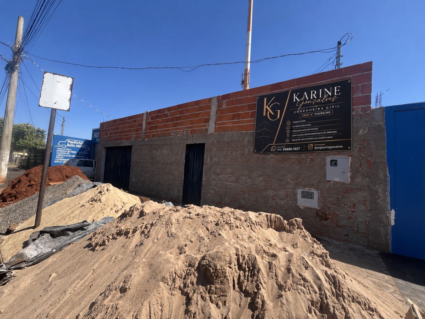 Fachada em alvenaria de tijolo aparente com placa de identificação da engenheira Karine Gonçalves e monte de areia no canteiro